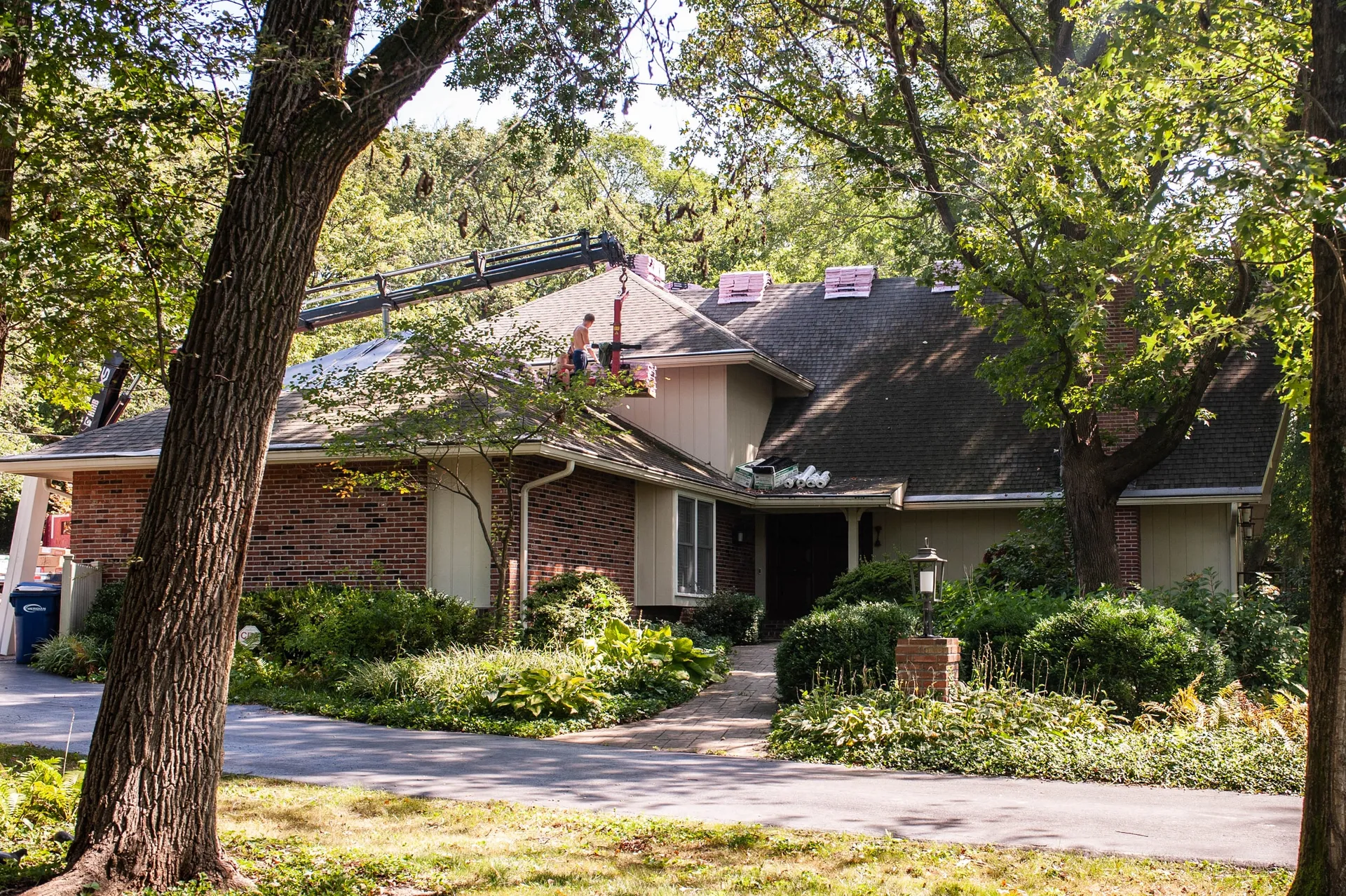 Roofing materials on a house roof, with a worker and delivery boom visible among trees