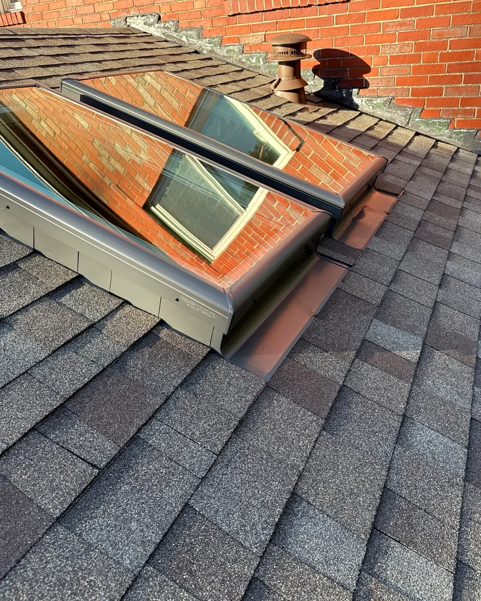 Two roof skylights integrated with shingles and flashing near a brick wall.